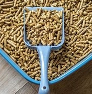 A tray of Sundown Kitt E Litter Scoop pellets with a grey plastic scoop on a wooden floor.